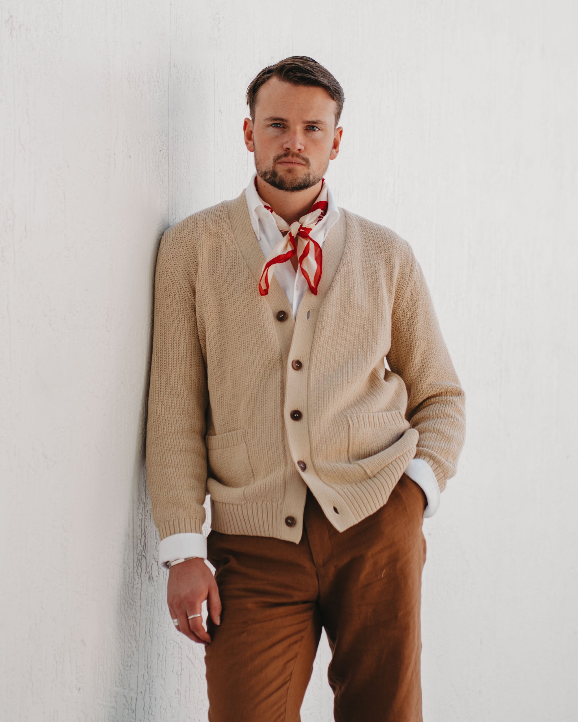 Man wearing a beige cardigan, white shirt, and brown pants with a red scarf against a white background